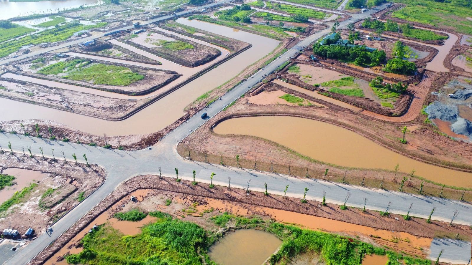 Ảnh chụp từ trên cao cho thấy mạng lưới đường nội khu và kênh đào tại dự án Alluvia City, với các ô đất bao quanh bởi dòng kênh uốn lượn, mặt đường đã trải nhựa, cây xanh được trồng đều hai bên đường; cảnh quan tổng thể đang dần hoàn thiện giữa khu vực đất nền còn đang thi công.