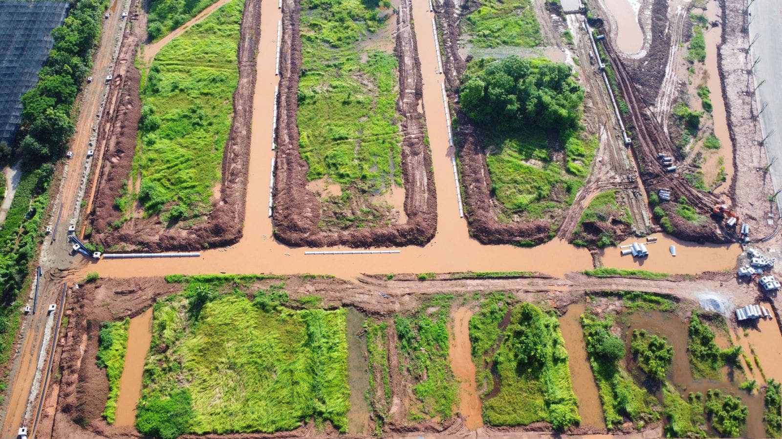 Ảnh chụp từ trên cao một khu vực đang thi công tại dự án Alluvia City, thể hiện các ô đất phân lô bao quanh bởi hệ thống kênh mương đã được đào, nước mưa đọng lại; xung quanh có máy móc thi công, vật liệu xây dựng, và các lối đi tạm trên nền đất đỏ ẩm ướt.